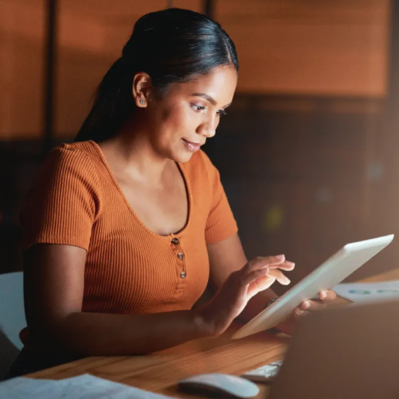 Woman in an orange top using a tablet at night in a modern office, surrounded by documents and softly lit workspace.