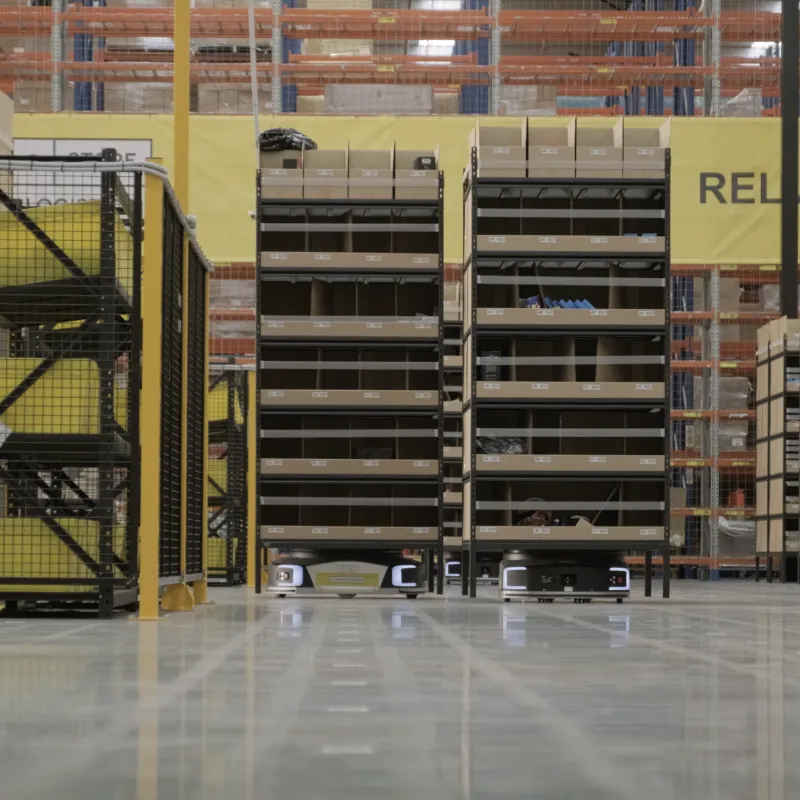 Automated warehouse with mobile robots transporting shelves, staff working on ladder, and organised storage racks in the background.